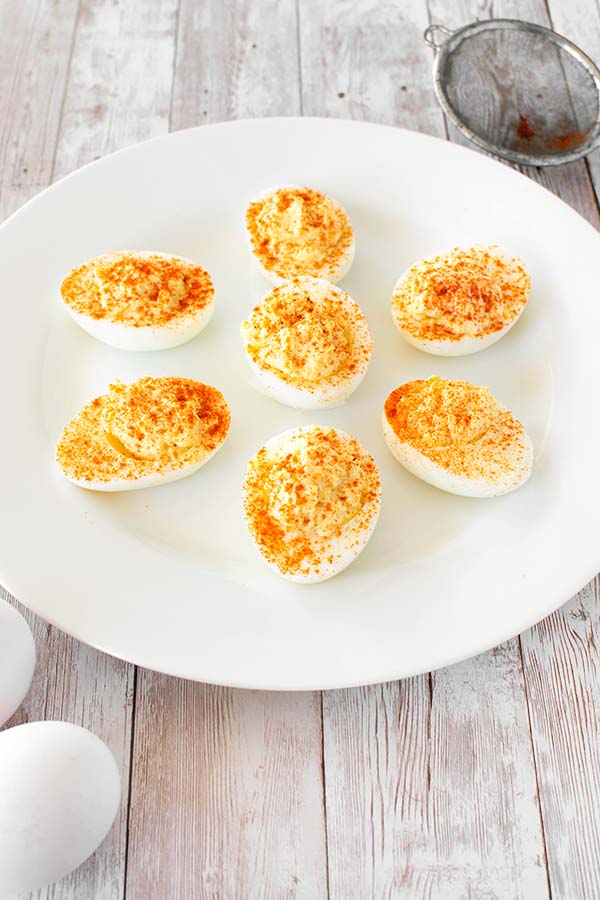 7 half deviled eggs on white plate sprinkled with paprika and two whole eggs with shell on the side