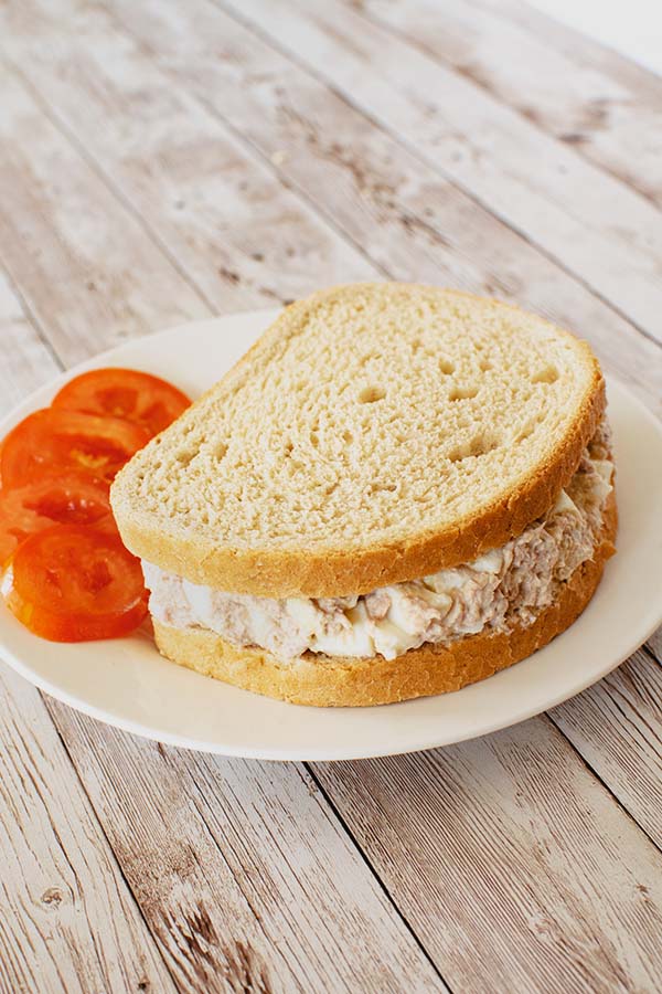 Tuna salad with hard boiled egg on seedless rye bread with sliced tomato on the side on a white plate