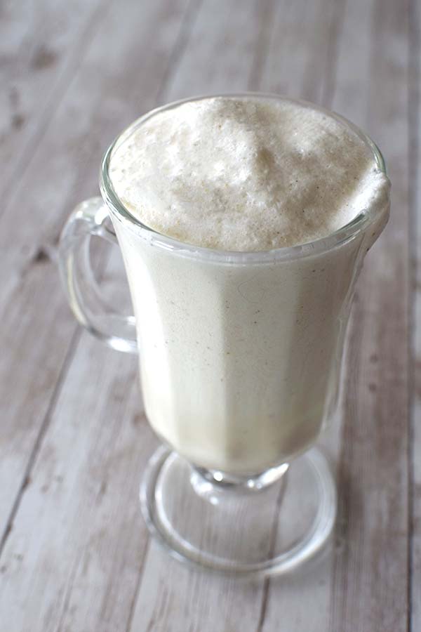 Eggnog with foam on top in a clear cappuccino glass on a white wood background