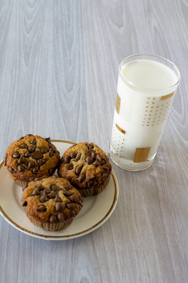 three banana chocolate chip muffins on a cream colored plate with gold trim with a glass of milk on a white wood background