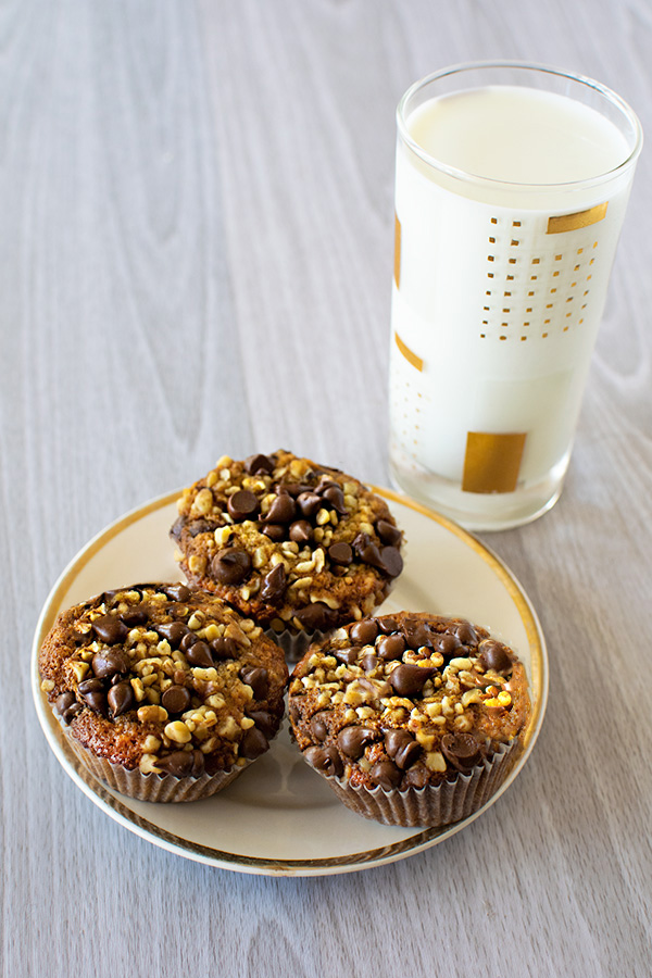 3 Banana Nut Chocolate Chip Muffins on a cream plate with a gold rim with a glass of milk on a white wood background.