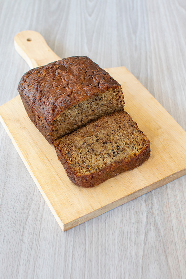 Banana bread loaf and a slice in from on a wooden cutting board on a white wood background