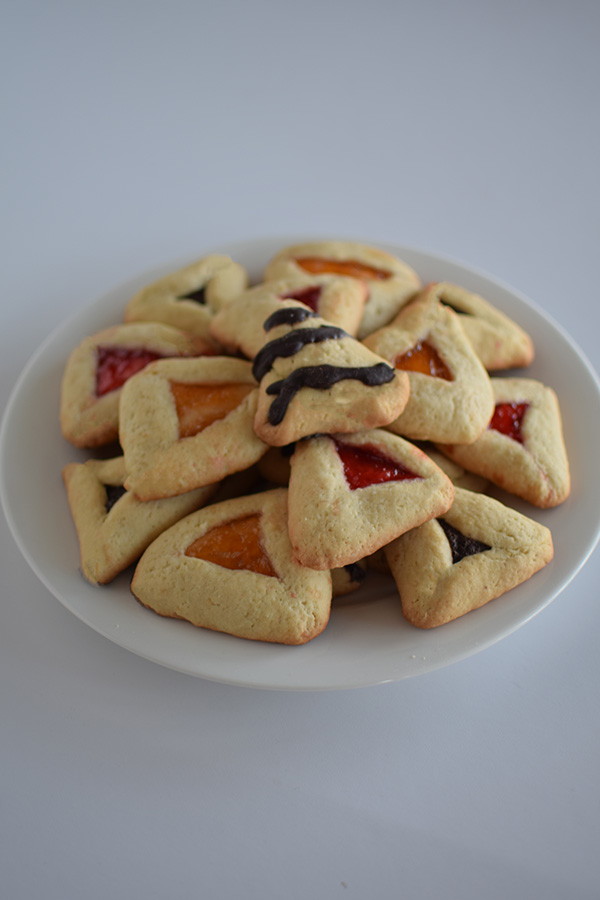 A plate full of hamentashen with a variety of fillings on a white background