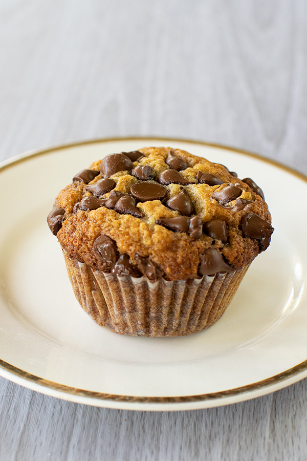 A single Banana nut chocolate chip muffin on a cream colored dish with a gold rim on a white wood background