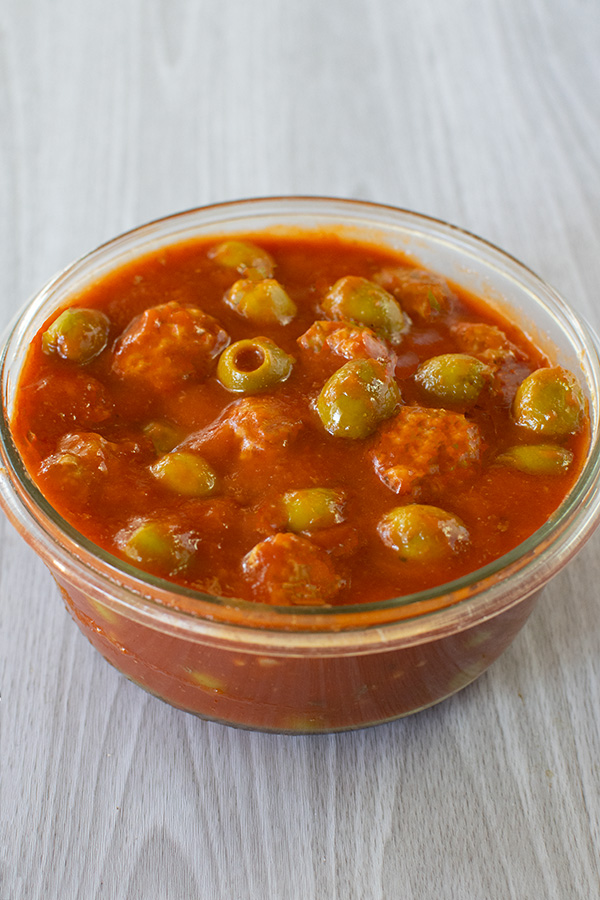 Olives and meatballs in a tomato sauce in a clear bowl on a white wood background
