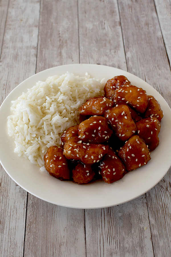 General Tso's Chicken covered with sprinkles of sesame seeds and rice on a white plate on a white wood background