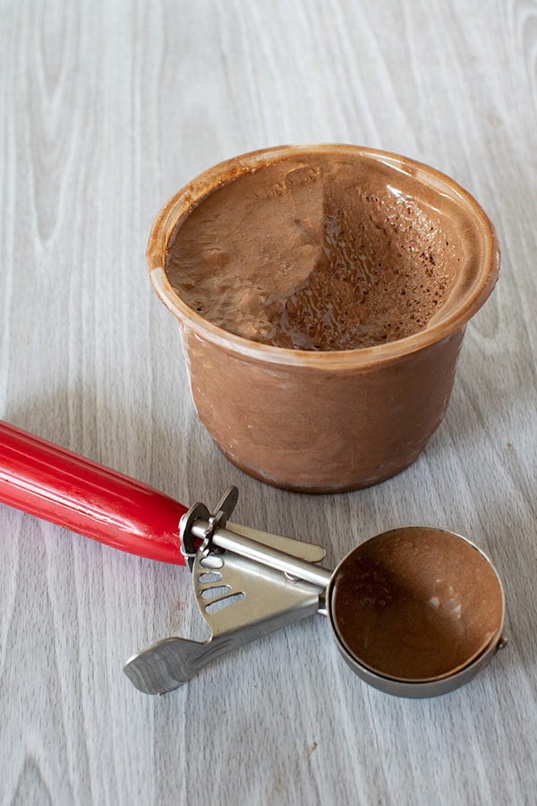 Small tub of chocolate ice cream with a scoop missing and a used ice cream scoop on a white wood background