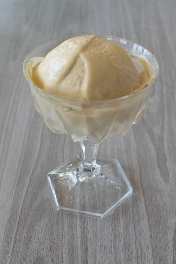 A large scoop of No-cook coffee ice cream in a clear glass dessert dish on a white wood background