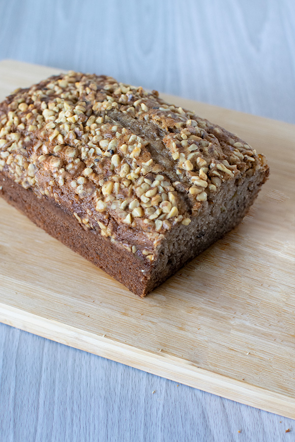 Banana nut bread loaf with a piece cut off on a wooden cutting board on a white wood background