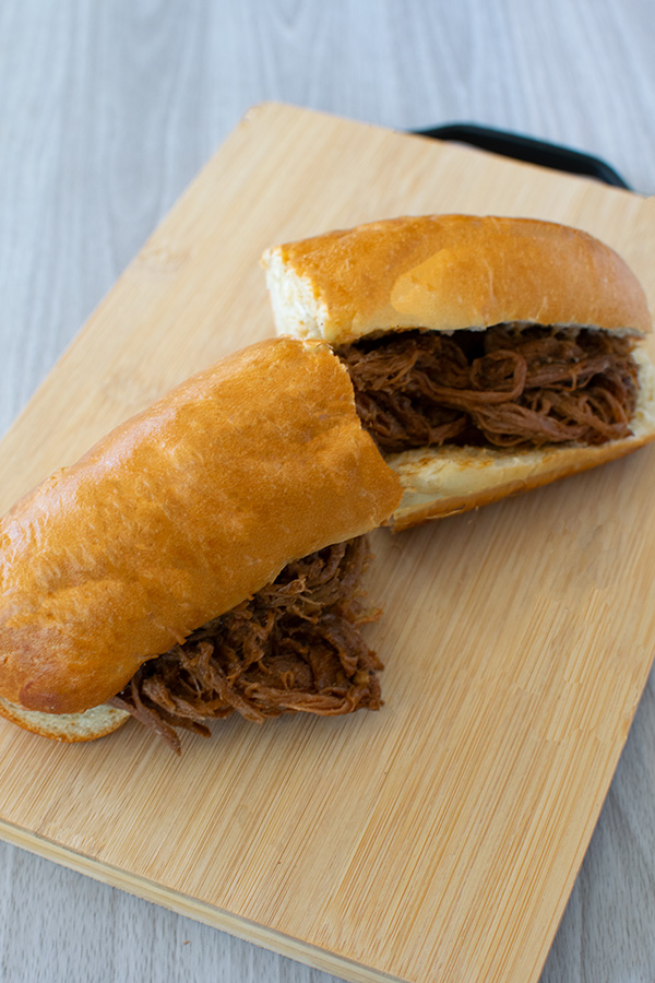 Pulled beef sandwich on a sub roll, cut in two, on wooden cutting board on white wood background