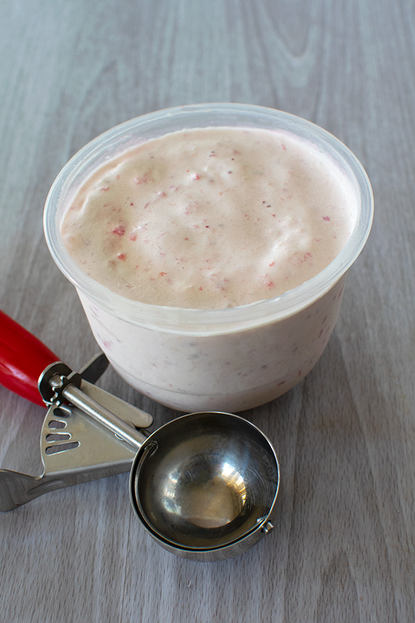 Strawberry ice cream in a container and a used ice cream scoop with a red handle on a white wood background