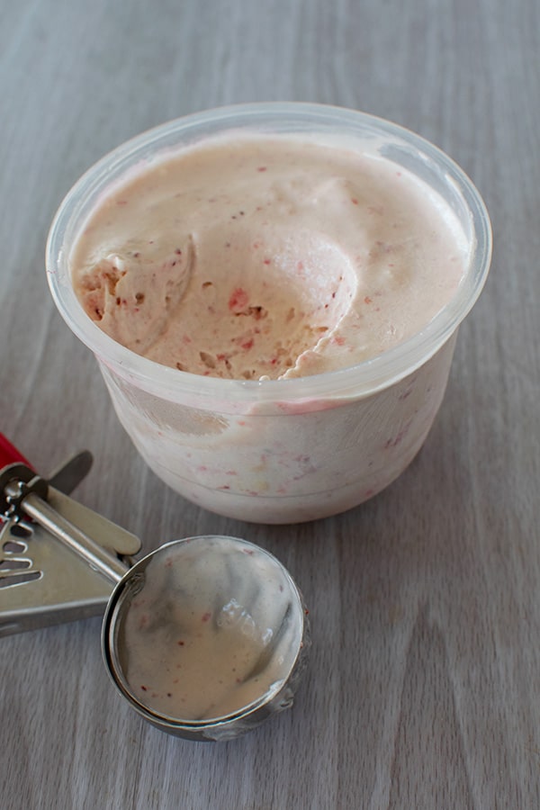 No-cook strawberry ice cream in a small tub and a scoop of ice cream missing and a used ice cream scoop in front on a white wood background