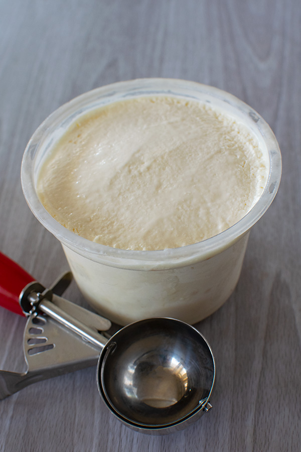 Vanilla ice cream in a container and a used ice cream scoop with a red handle on a white wood background