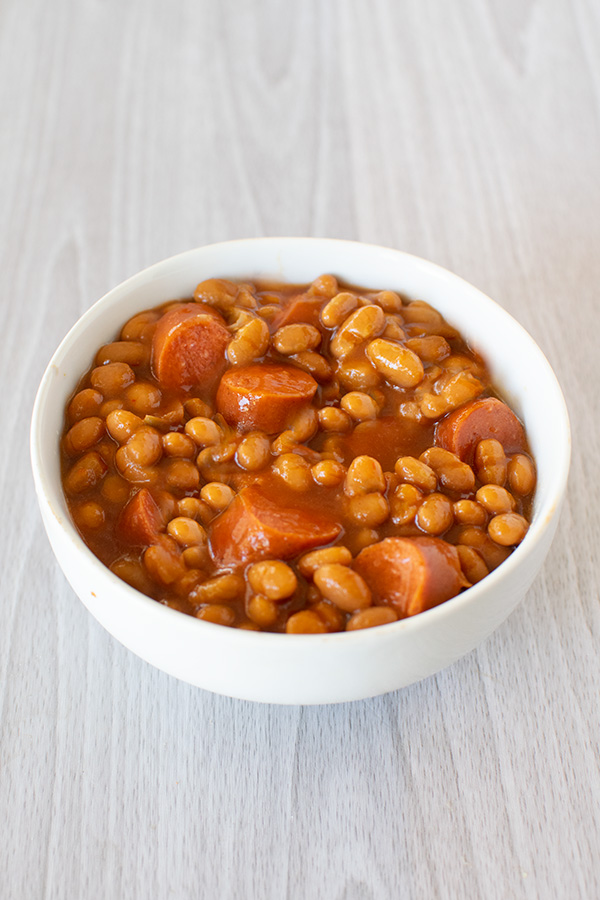 Baked beans and hotdog slices in a white bowl on a white wood background