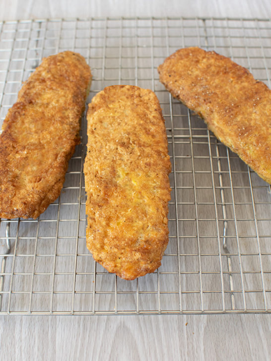 Three breaded and fried zuccini cutlets on a wire cooling rack on a white wood background