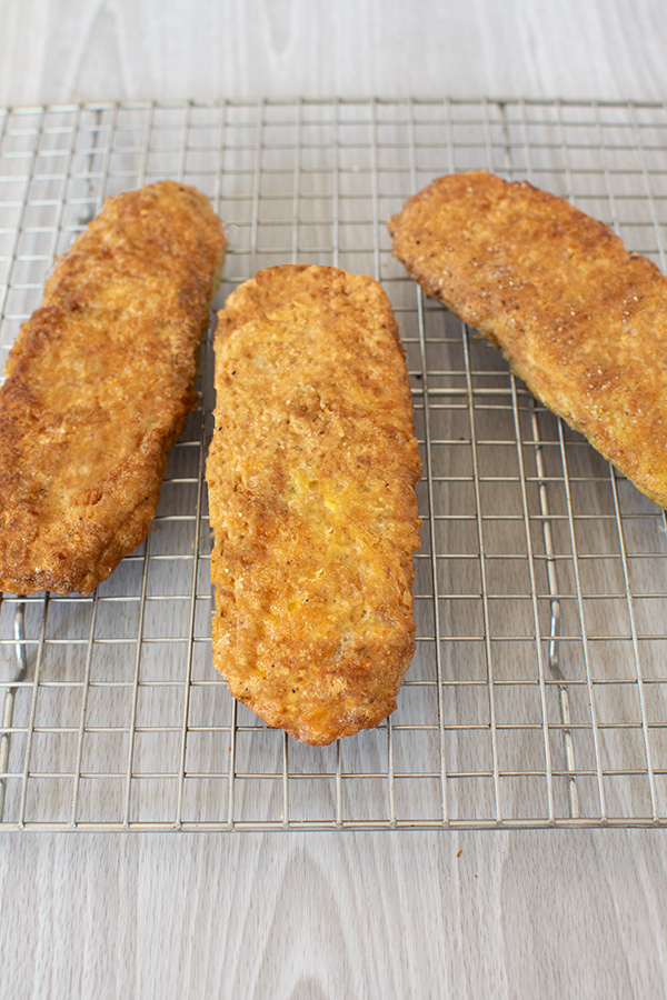 Three breaded and fried zuccini cutlets on a wire cooling rack on a white wood background