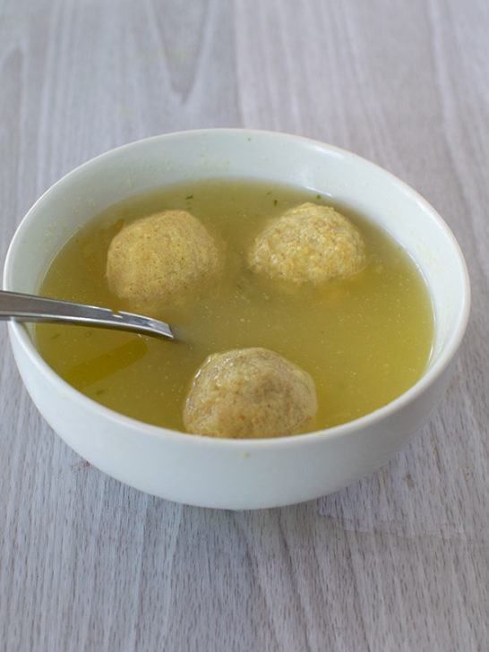 3 matzo balls in chicken soup in a white bowl with a spoon on a white wood background
