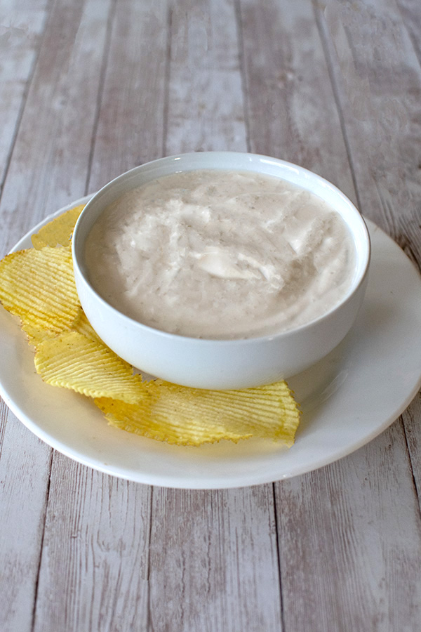 Onion dip in a white bowl on a white plate with potato chips on a white wood background