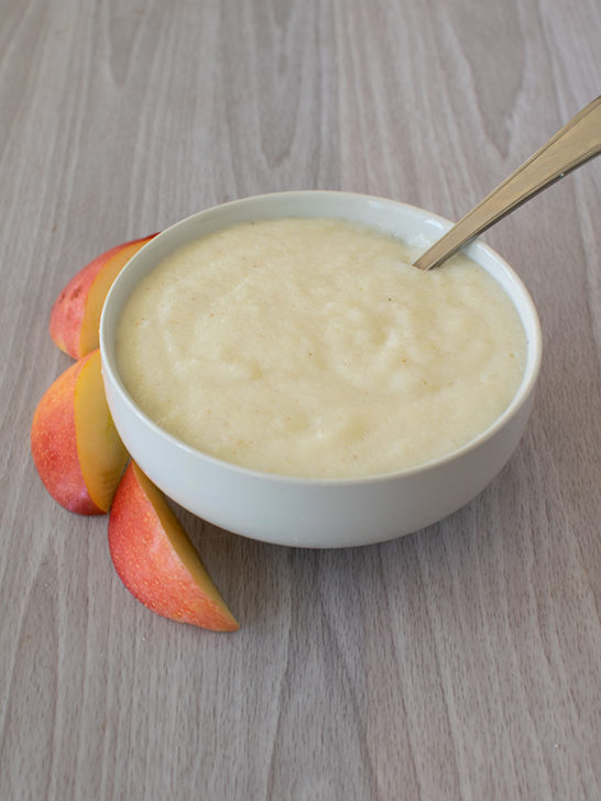 A white bowl of dairy free farina with a spoon and sliced peach on the side on a white wood table.