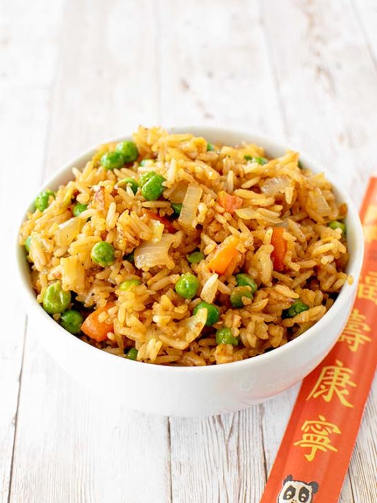 Vegetarian fried rice with peas, carrots, and onions near a package of chop sticks on a white wood table