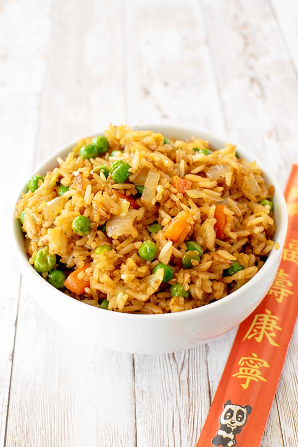 Vegetarian fried rice with peas, carrots, and onions near a package of chop sticks on a white wood table