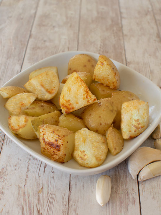Vegetarian Roasted Garlic potatoes on a square white plate on a white table with garlic cloves nearby