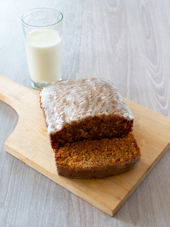Honey Loaf Cake with Glaze with a slice off on a wooden cutting board with a clear glass of milk nearby on a white wood table.