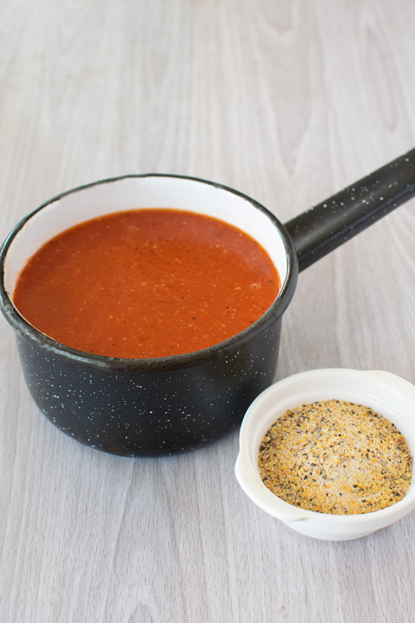 A saucepan with vegetarian pasta sauce and a bowl of the spices on a white wood table