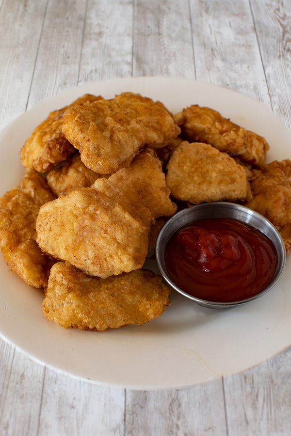 Cornflake chicken nuggets with a little metal bowl on a white plate on a white wood table