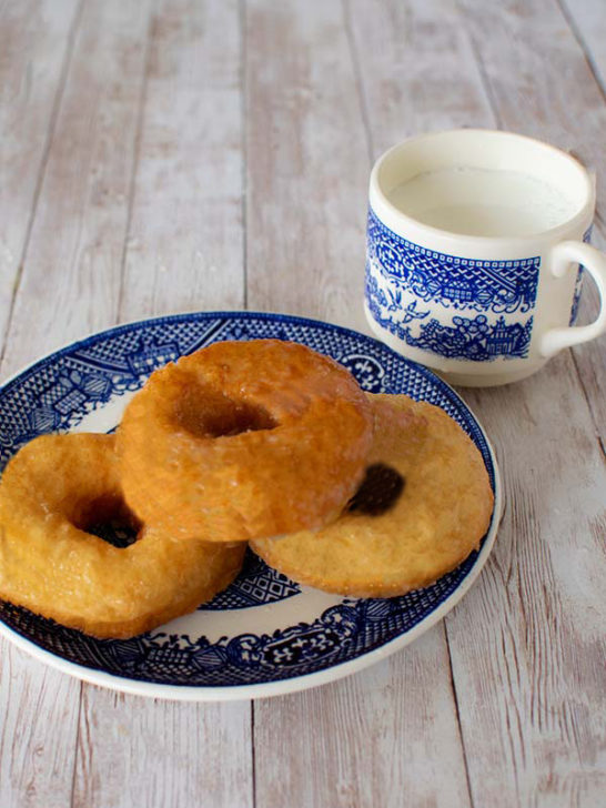 Three plain dairy free doughnuts on a blue and white plate with blue and white cup of milk nearby on a white slatted wood table
