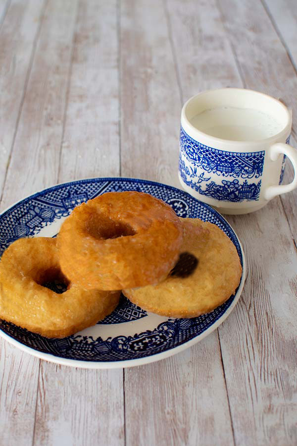 Three plain dairy free doughnuts on a blue and white plate with blue and white cup of milk nearby on a white slatted wood table