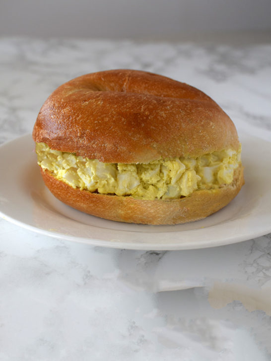 Egg salad on a bagel on a white plate on a white marble background