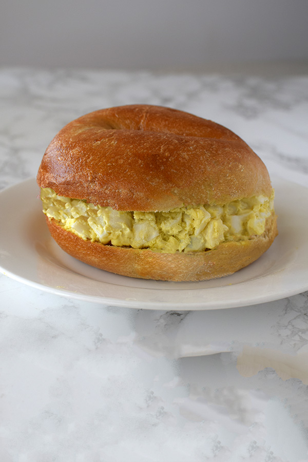 Egg salad on a bagel on a white plate on a white marble background