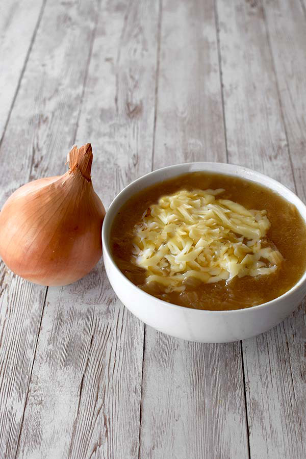 French Style Onion Soup topped with grated cheese in a white bowl near an onion on a white wood table