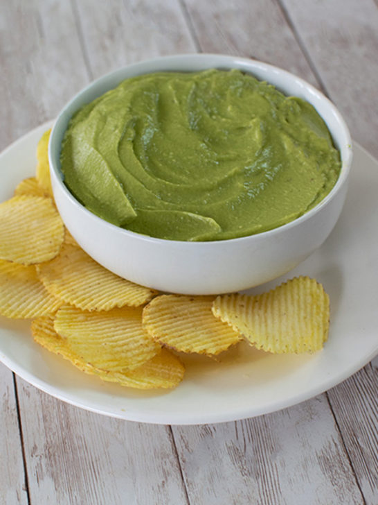 Avocado Dip in a white bowl on a white plate with potato chips on a white wood table