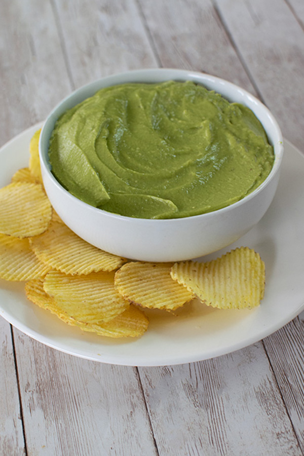 Avocado Dip in a white bowl on a white plate with potato chips on a white wood table
