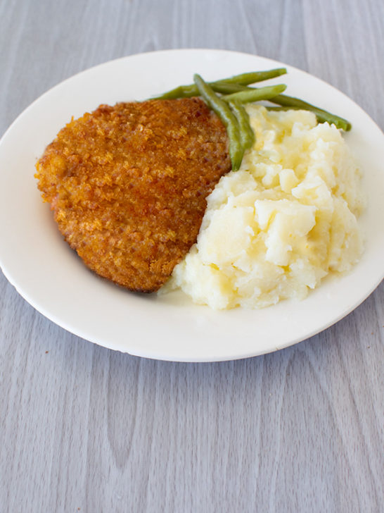 Cornflake Fried Chicken on a white plate with green beans and mashed potatoes on a white wood table
