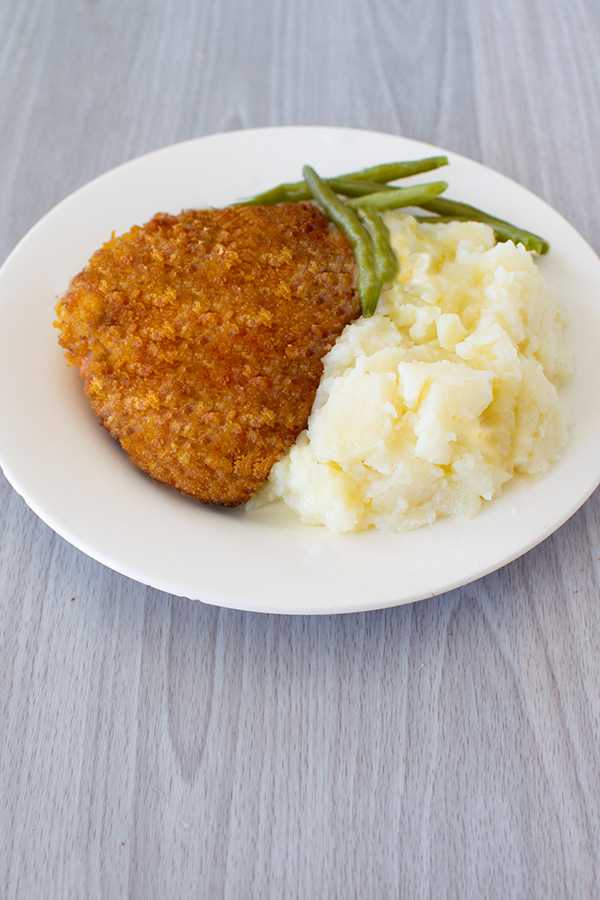 Cornflake Fried Chicken on a white plate with green beans and mashed potatoes on a white wood table