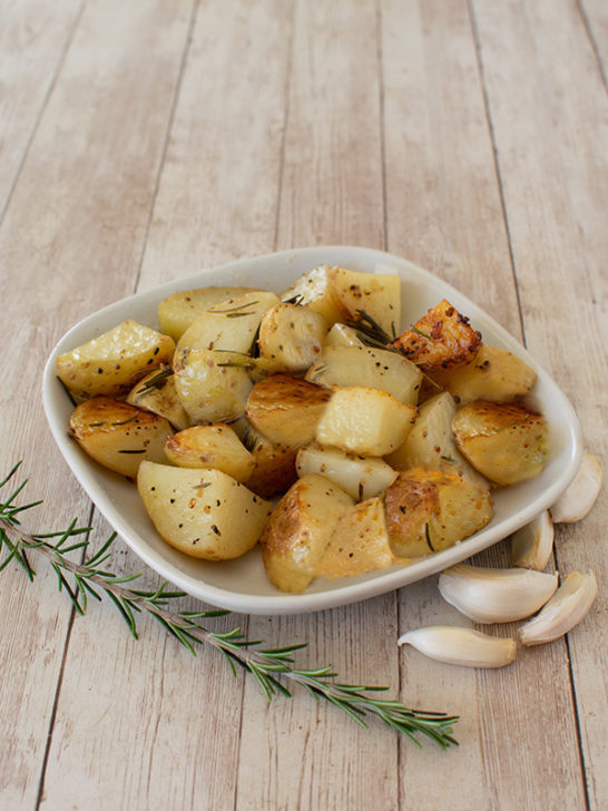 Garlic and Rosemary Potatoes on a white square plate on a white wood table with garlic cloves nearby