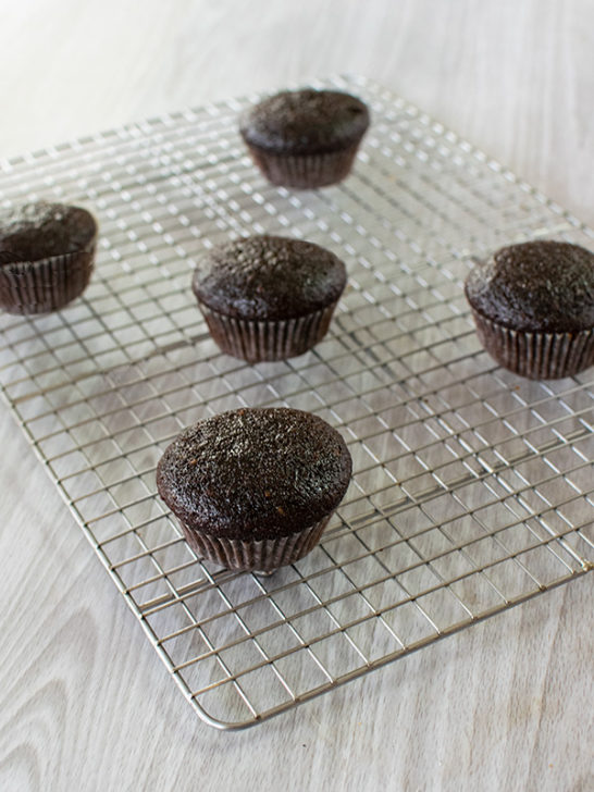 Dairy free chocolate cupcakes and muffins on a drying rack on a white wood table.