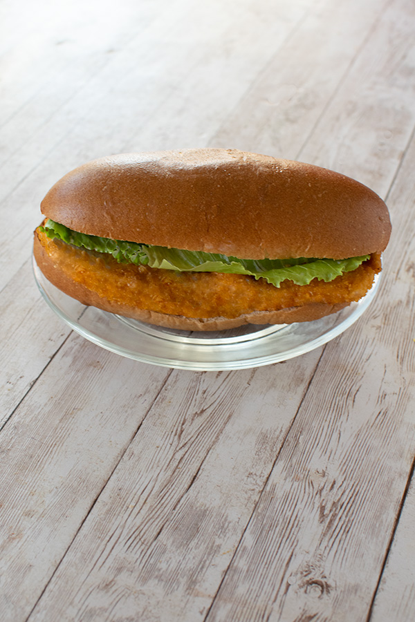 Schnitzel sandwich with a whole wheat roll and lettuce on a white plate on a white wood table