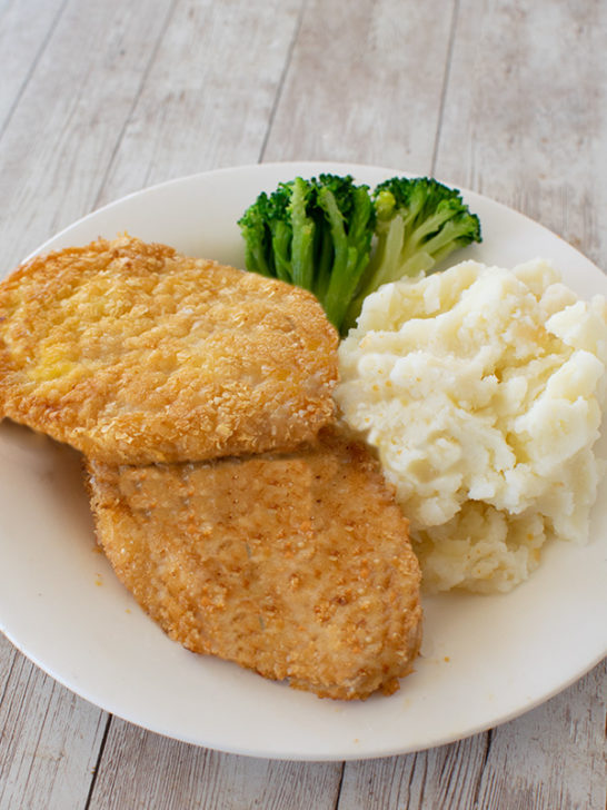 Two potato flake schnitzels for passover on a white plate with mashed potatoes and broccoli on a white wood table
