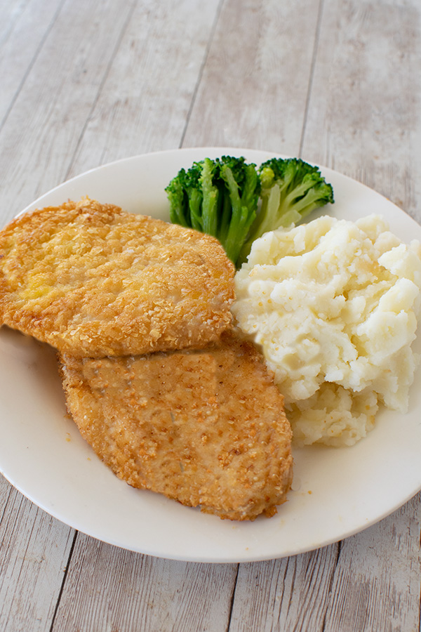 Two potato flake schnitzels for passover on a white plate with mashed potatoes and broccoli on a white wood table