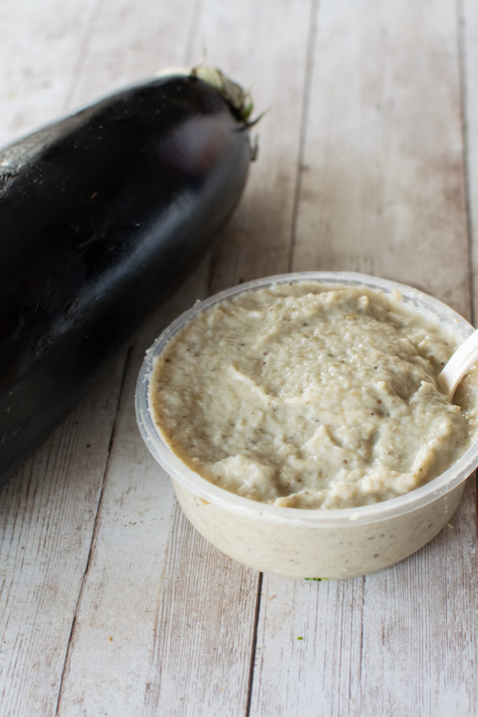Eggplant salad with tahini in a container with a spoon near a raw eggplant on a white wood table.