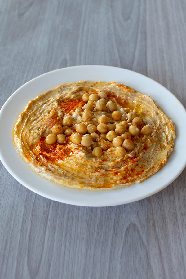A white plate of chumus with chickpeas and paprika on a white wood table