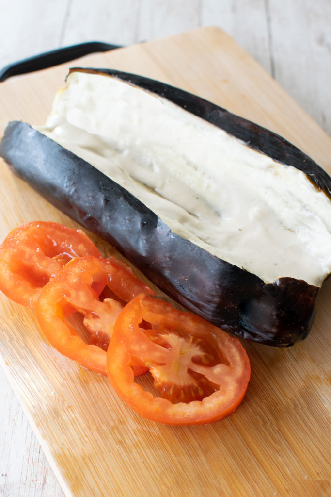 Roasted eggplant sliced open with tehini poured on near slices of tomato, all on a wooden cutting board.