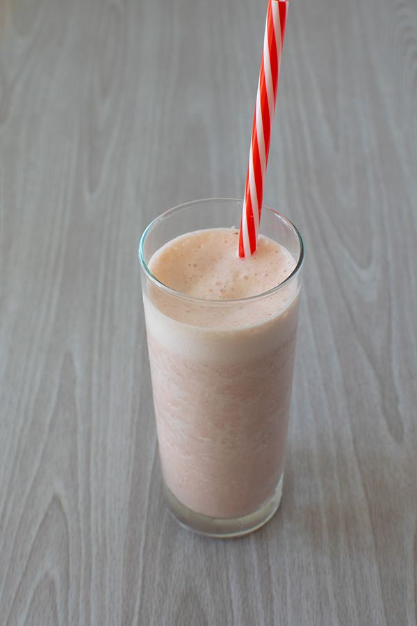 Pink melon smoothie in a clear glass with a red and white straw on a white wood table
