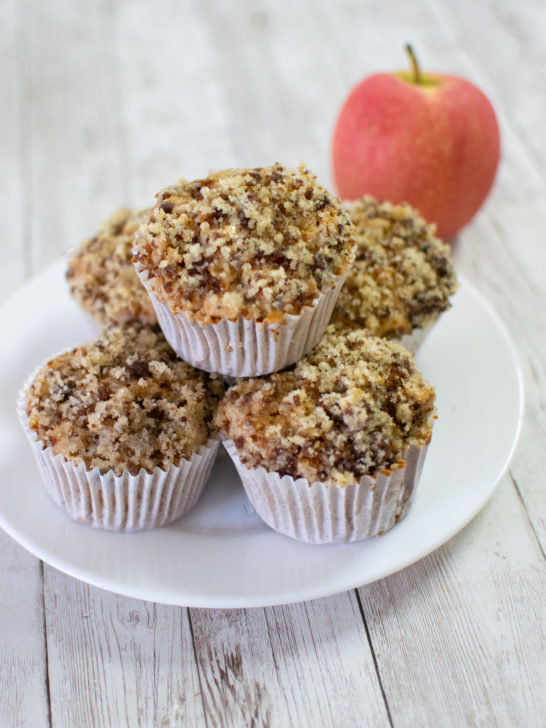 Easy Dairy-Free Apple Cinnamon Muffins piled on a white plate next to a red apple