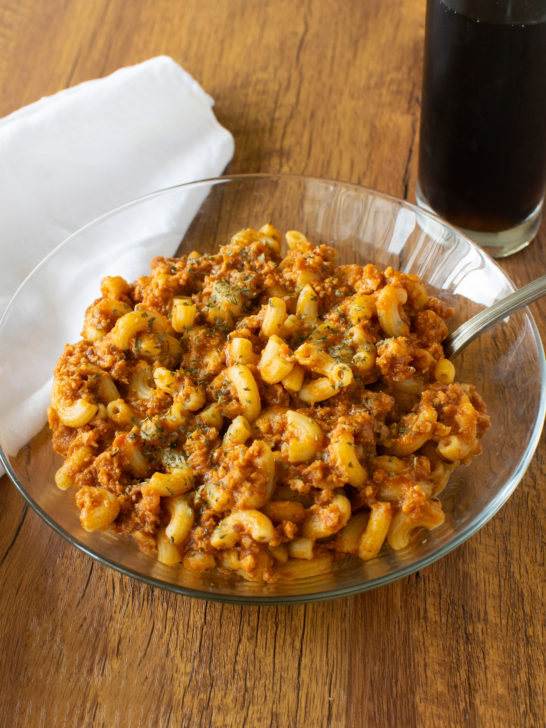 Easy American Goulash in a clear bowl with a fork on a dark wood table with a white napkin to the left and a clear glass of coke on the right