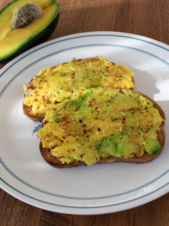 2 slices of Really easy avocado toast on a white place with half and avocado nearby on a dark wood table.